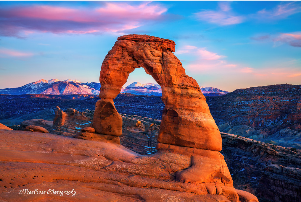 Delicate Arch in Arches National Park, Moab, Utah at sunset with snow-capped La Sal Mountains in the background, dramatic red rock desert landscape photograph.
