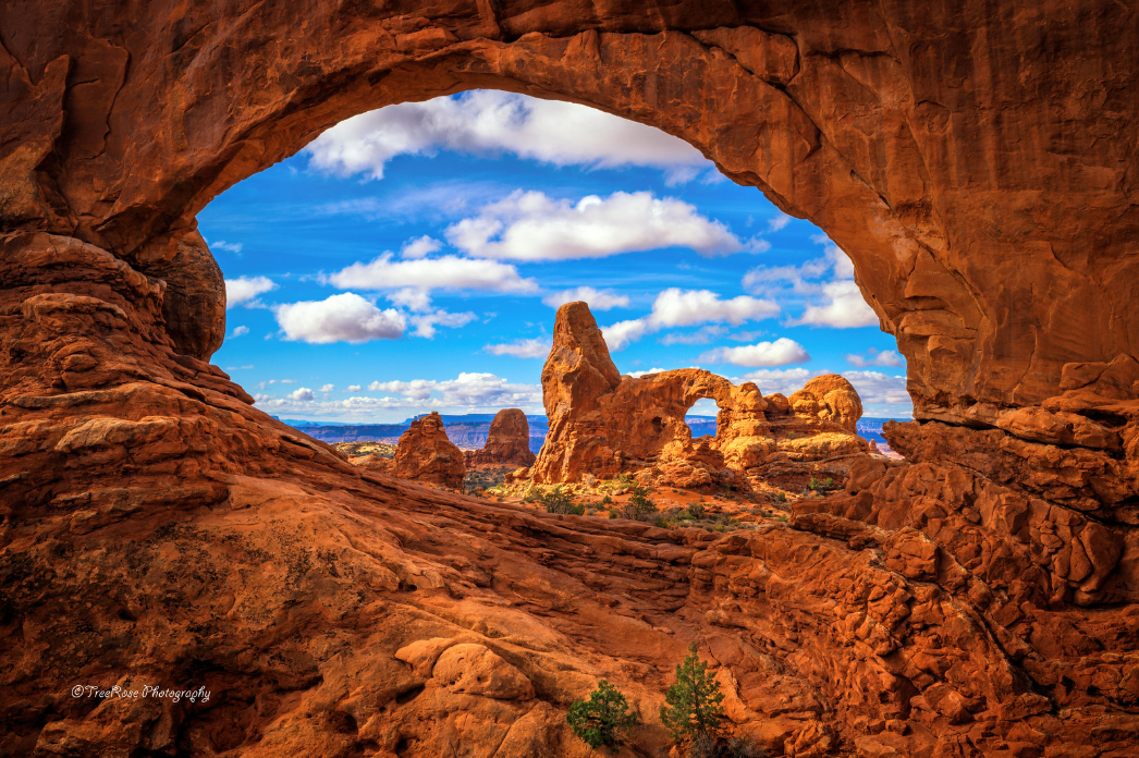 View of Turret Arch through North Window Arch in Arches National Park, Moab, Utah with dramatic red rock formations under a bright blue sky and scattered clouds.